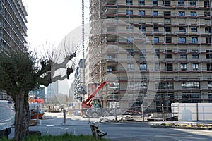 Construction site of an apartment house, rough building covered by scaffolding and protective net. Lateral view with a tree