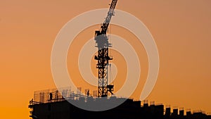 The construction site of a large apartment building. Silhouettes of workers and cranes on the construction site against the sunset