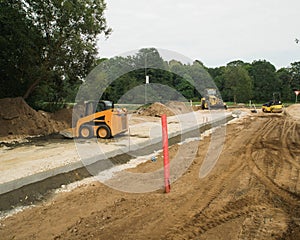 Construction site of new road in suburban. Yellow excavator, road roller and skid loader. Green trees.