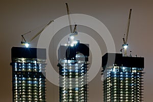 Construction of three high-rise buildings using cranes. Night construction of skyscrapers with floodlights