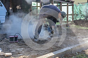 A construction worker cuts a concrete curb using a professional angle grinder causing a cloud of dust and smoke