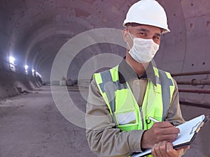 Construction worker dressed in medical mask, uniform and white hardhat stands with clipboard at metro tunnel