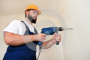 Construction worker, handyman and builder with blue electric drill drills a nail hole into wall of construction site.