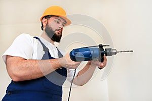 Construction worker, handyman and builder with blue electric drill drills a nail hole into wall of construction site.