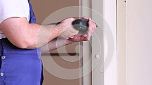 Construction worker and handyman with electric drill drills nail hole into wooden wall door at construction site.