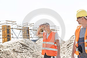 Construction worker looking at tired colleague wiping sweat at site