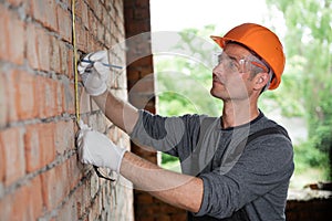 construction worker male foreman in protective helmet and goggles measures the brick wall ruller. Repair and