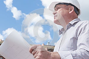 A construction worker man in white helmet holding blueprints on a background with blue sky