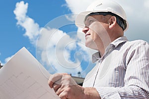 A construction worker man in white helmet holding blueprints on a background with blue sky