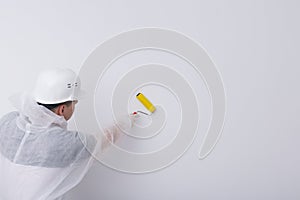 A construction worker in protective clothing and a helmet, smoothes the wallpaper on the white wall with a special roller, there