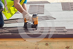 Construction worker putting the asphalt roofing shingles with nail gun on a new frame house.