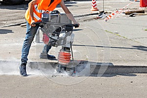 A construction worker is ramming a trench with a vibrating rammer at a construction site and kicking up a cloud of dust around him