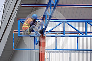 Construction worker with safety equipment welding metal on roof structure of warehouse building in construction site