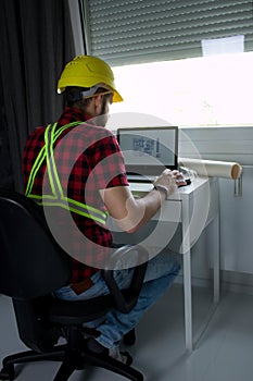 Construction Worker is sitting at his desk at the computer, looking at building plans, counting and thinking