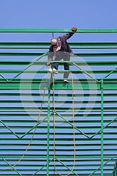 Construction worker is welding metal on green roof warehouse structure against blue sky background