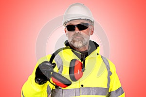 A construction worker in a white hat,  yellow hi-viz coat and dark tinted safety glasses on the light red background gives foam-