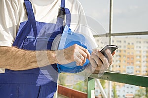 Construction worker in a work attire holds a construction helmet, mobile phone and dial number in hand. Work at high altitude. S