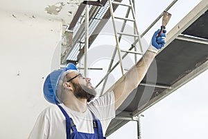 Construction worker in a work attire, protective gloves and a helmet on the head gives a spatula. Work at high altitude. Scaffol