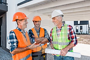 Constructors in helmets standing near coworker with blueprint