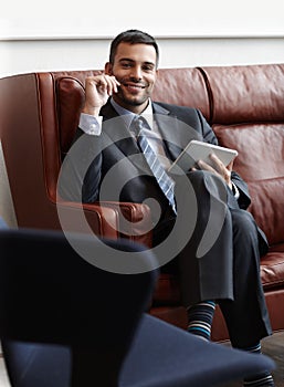 The consummate businessman. A smiling young businessman sitting on a leather sofa while using a digital tablet.