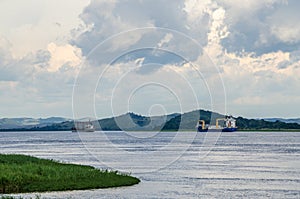 Container cargo ships on mighty Congo river with dramatic cloudy sky and lush green grass in foreground, DRC, Africa