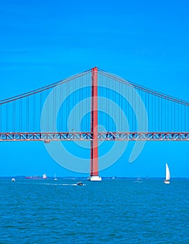 Container ship, motorboats and sailboats navigating the Tagus River crossing the red steel 25 de Abril suspension bridge with road