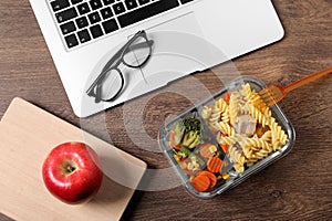 Container of tasty food, laptop, apple, book and glasses on wooden table, flat lay. Business lunch