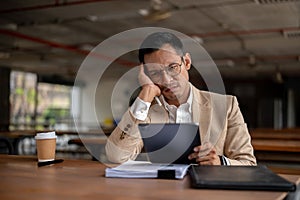 A contemplative Asian businessman looks pensive while holding a tablet, facing a problem at work