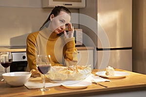 A contemplative woman sits at a table with a slice of cake and a glass of red wine in a warmly lit kitchen. A woman is