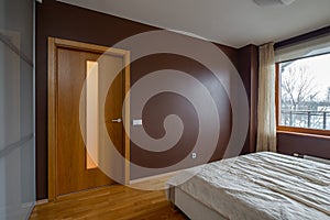Contemporary interior of bedroom in modern flat. Cozy bed. Huge window. Hardwood floor. Wooden door.