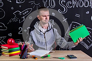 Contempt youngster sitting at the desk with a pile of books in the hand, surrounded with school supplies. Chalkboard as