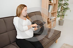 Content pregnant woman holding her tablet sitting on a couch in the living room at home