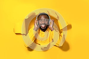 Contented african american man peeking out hole on yellow background and listening to music in headphones