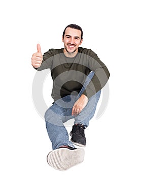 Contented casual young man sitting on the floor smiles to camera, showing thumb up approval gesture. Lifestyle full length