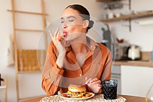 Contented caucasian lady licking her fingers while eating delicious french fries and hamburger, kitchen interior