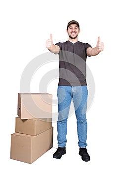 Contented delivery man wearing cap, full length portrait, shows thumbs up, stands near the parcel post boxes, isolated on white.