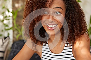 Contented elated African Amerian woman has crisp hair, looks with toothy smile aside, wears striped summer t shirt