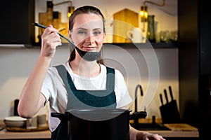Contented, happy young housewife woman in an apron holds a spoon of a pot of soup, tastes soup from a pot. The concept