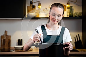 Contented, happy young housewife woman in an apron holds a spoon of a pot of soup, tastes soup from a pot. The concept