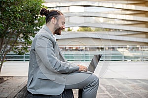 Contented modern businessman with topknot working on laptop outdoors