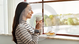 Contented young woman enjoying a peaceful moment, sipping coffee and gazing out a sunny window