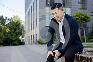 Contorted in pain, a young Asian businessman sits on a bench near an office center and holds his knee with his hands