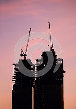Contours of tower cranes above the high constructing tower buildings against evening sky in the red sunset