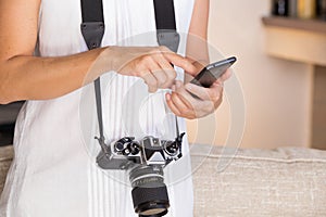 Contrast between old and modern times: a young woman with a vintage camera around her neck fiddles with her smartphone photo