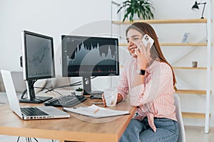 Conversation by the phone. Female stockbroker in casual clothes is working in the office by pc