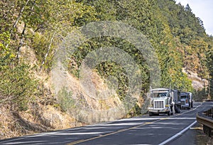 Convoy of the big rig tipper semi trucks with tip trailers running on the mountain winding road with trees on the hills