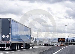 Convoy of big rigs semi trucks with different semi trailers running on the interstate highway with bridge and road sign electronic