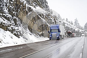 Convoy of the big rigs semi trucks with semi trailers standing on the wet road shoulder on mountain pass at winter snowy weather