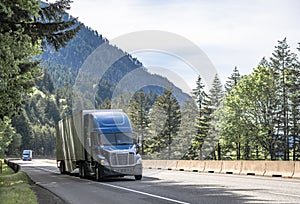 Convoy of blue big rigs semi trucks with semi trailers transporting cargo driving on the turning wide highway road with green