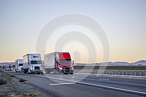 Convoy of industrial big rigs semi trucks with semi trailers driving with cargo on the straight highway road at twilight time